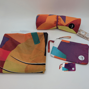 A folded up section of brightly colored polyester banner offcuts next to a Morph Bags Essentials Pouch and a rolled up Yoga Mat Bag made from the same material, illustrating the transformation from waste to finished product.
